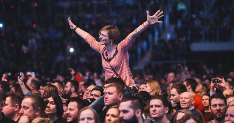 Event venue full of people looking at and listening to an event. One woman sitting on someone's shoulders, enthusiastically spreading her arms, cheering