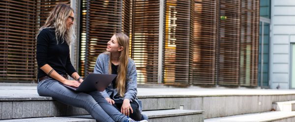Woman with laptop speaking to another woman, sitting on the stairs in front of public educational building